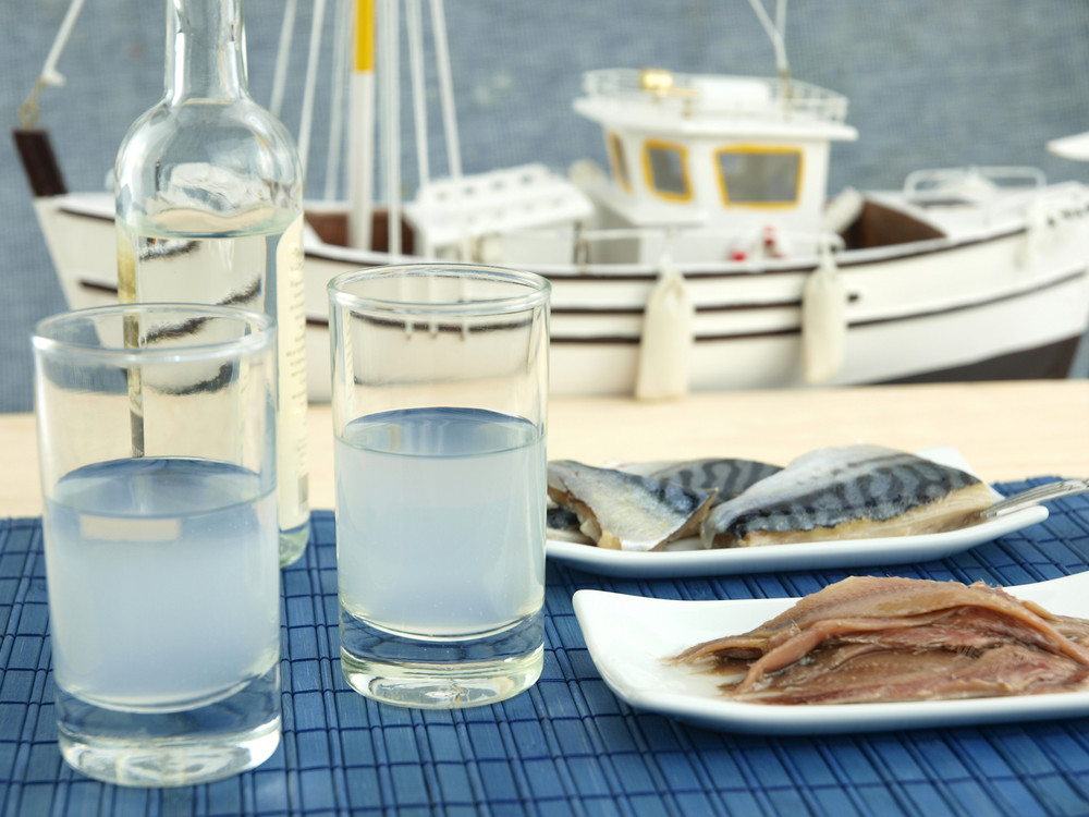 Tisch im Freien m. 2 Gläsern Ouzo u. Fisch, im Hintergrund ein Fischerboot.