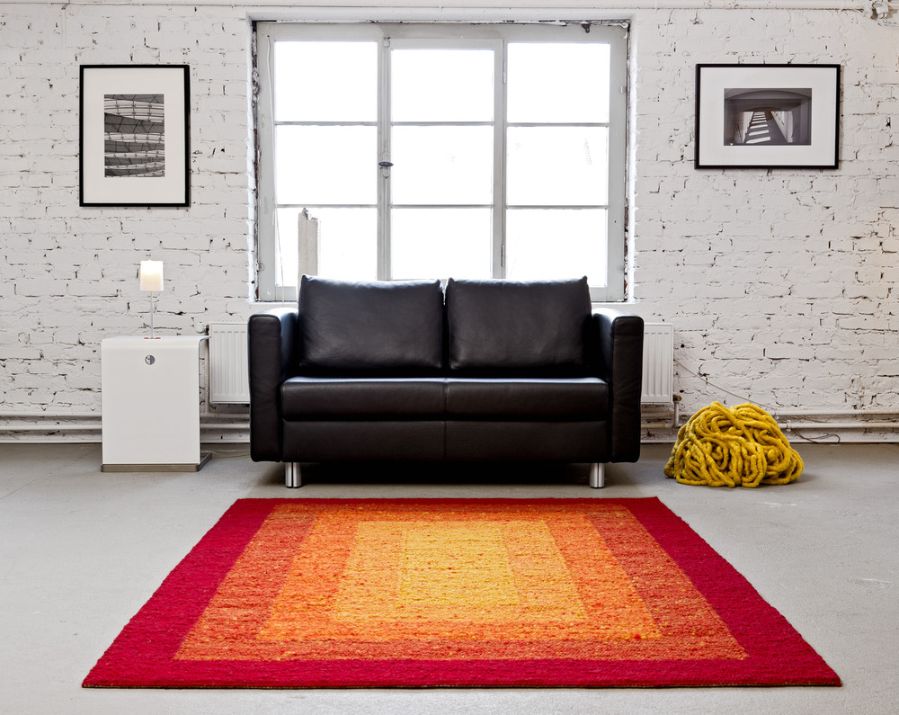 Gelb-orange-roter Teppich vor schwarzem Sofa in hellem Zimmer. Foto: Habbishaw Teppiche 